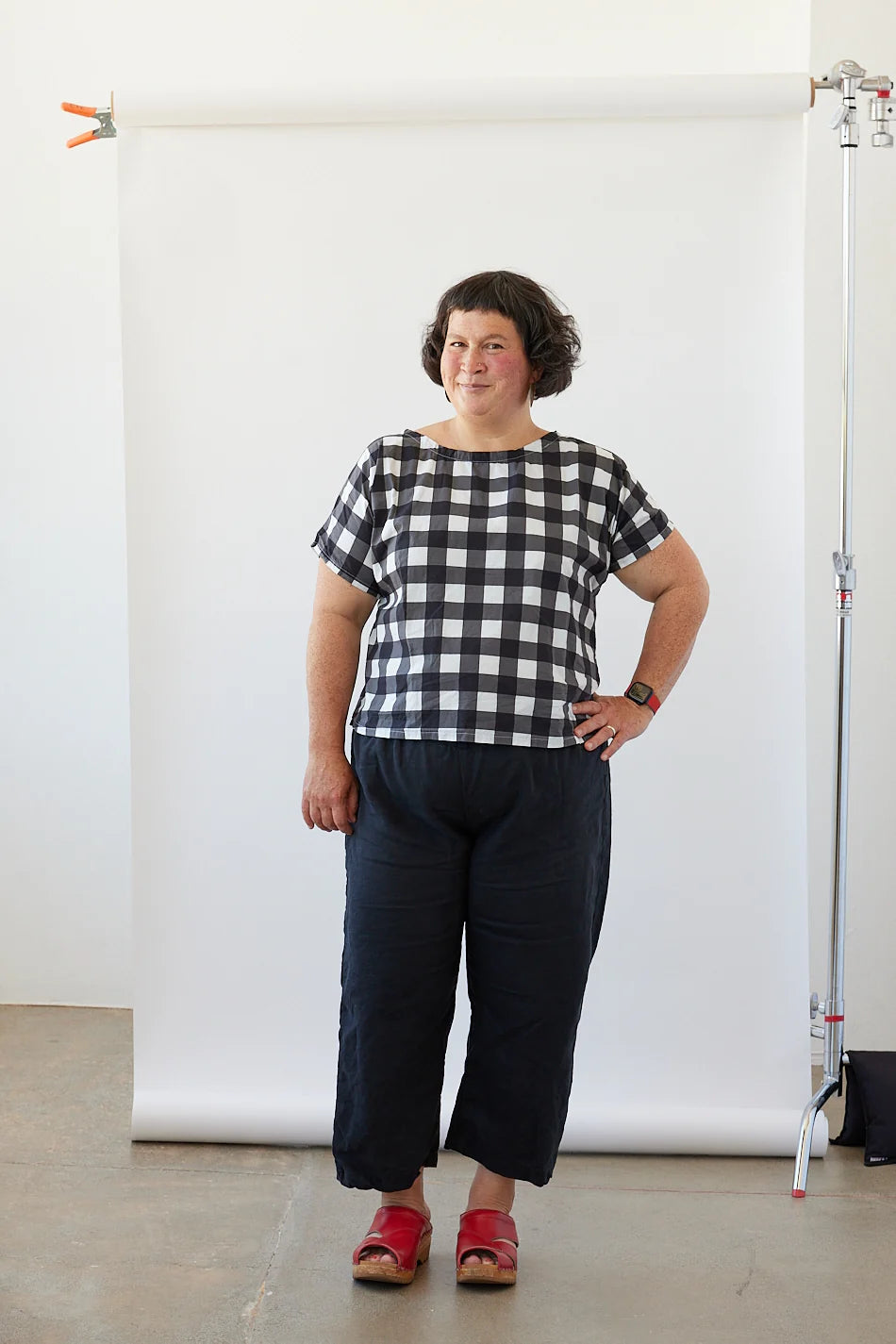 Person wearing a black and white checkered shirt and dark pants standing against a white backdrop.