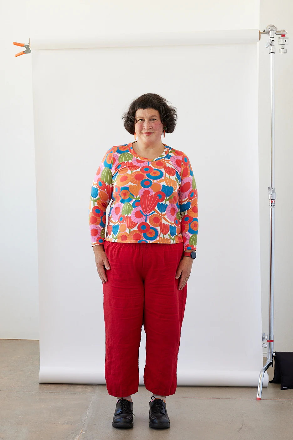 Person wearing a colorful floral top and red pants standing in front of a white backdrop.