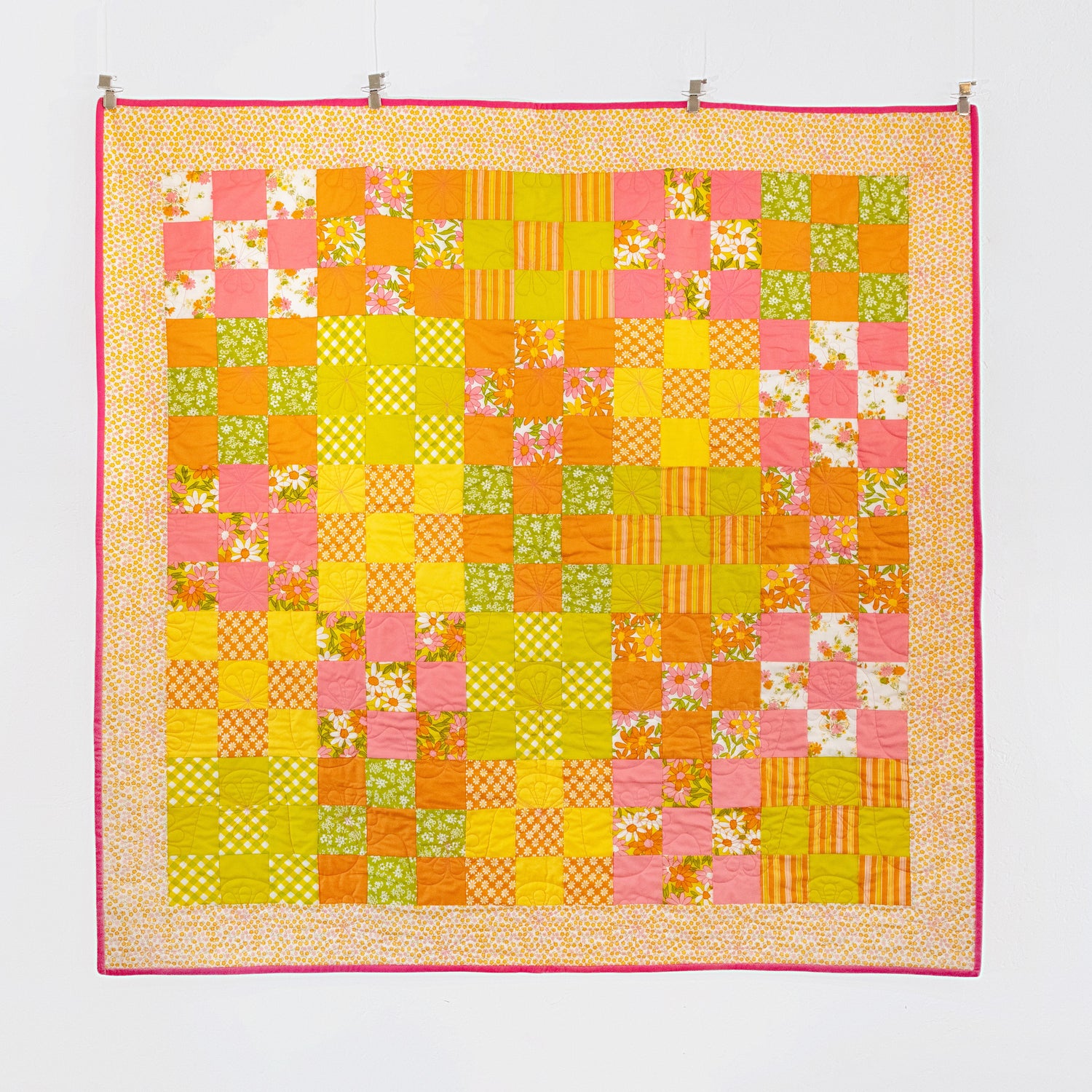 A patchwork quilt hanging on a white wall, featuring orange, yellow, pink and white fabrics arranged in a checkerboard pattern