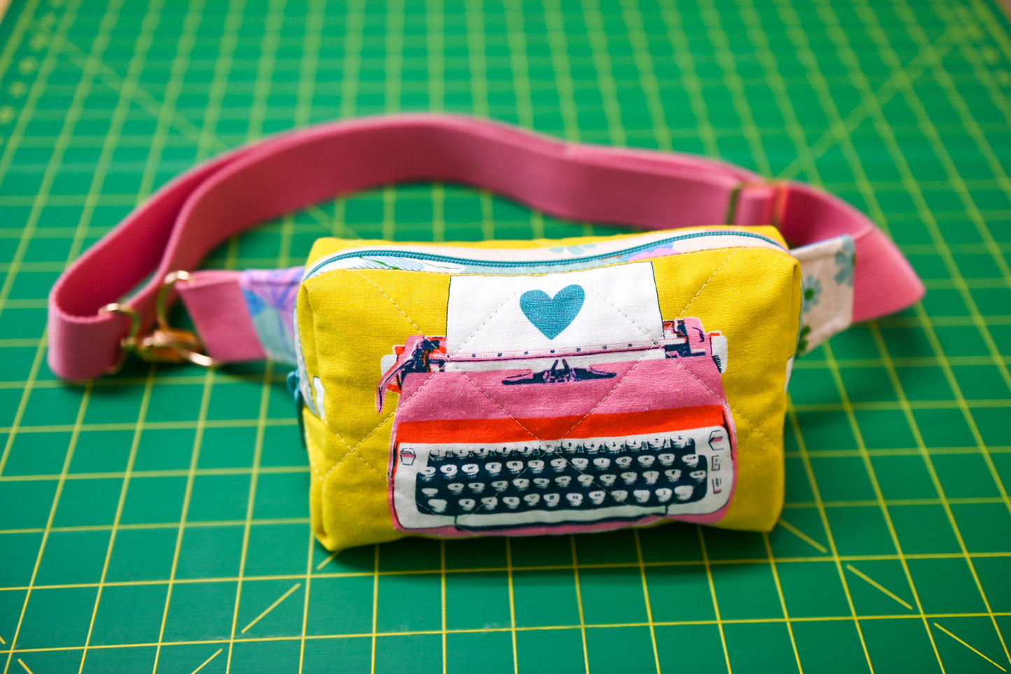 Yellow pouch with a pink typewriter design on a pink belt on a green cutting mat.