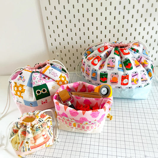 Four fabric drawstring pouches in assorted colorful prints and patterns, arranged on a white grid mat against a white pegboard background.