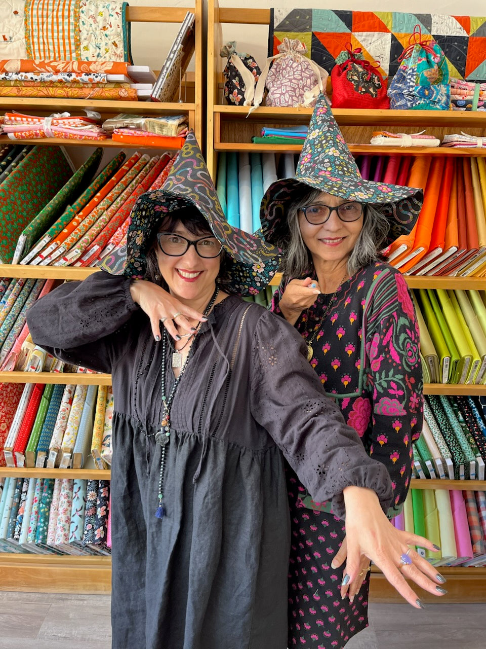 Two women wearing witch hats and patterned dresses posing in front of shelves filled with colorful fabrics in a shop.
