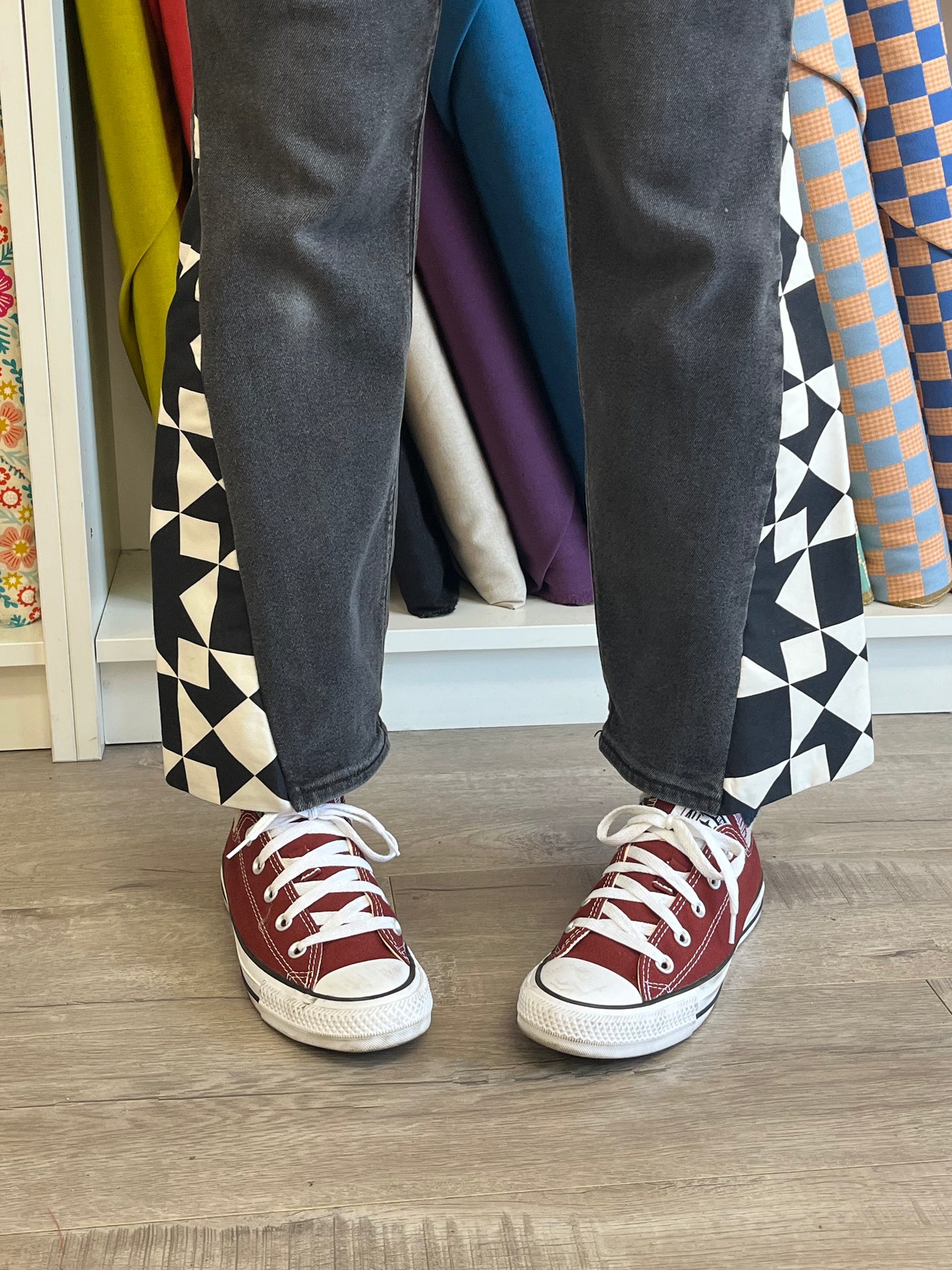 Person wearing maroon sneakers with white laces on a wooden floor, with colorful fabric rolls in the background.