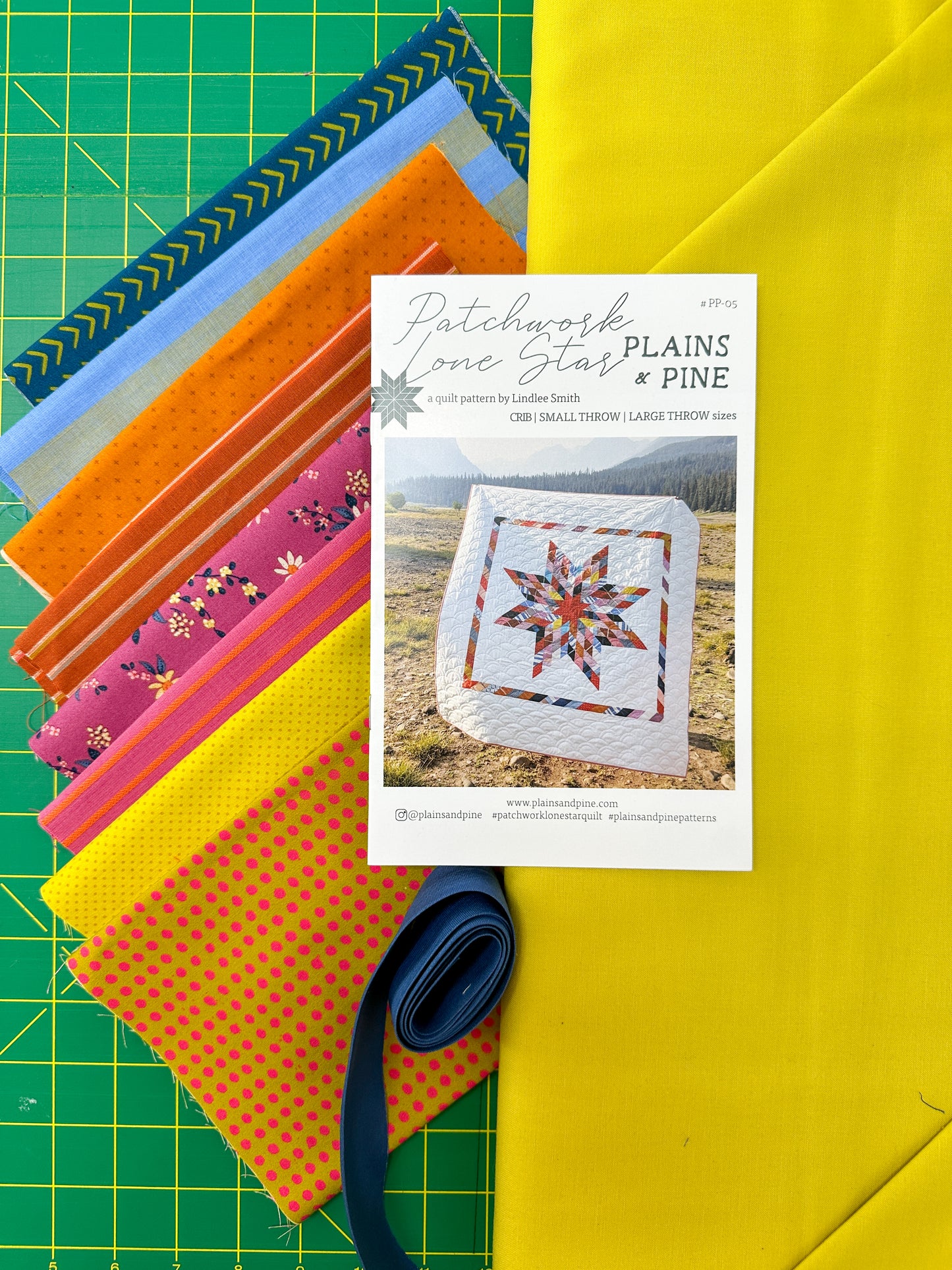 Colorful fabric strips on a green cutting mat with a quilt pattern book titled 'Plains Pine'.