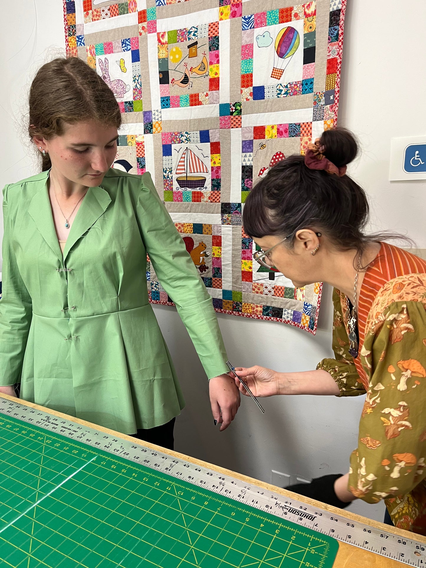 A woman in a light green peplum-style jacket or dress stands at a table while another woman measures her arm with a tape measure. A green cutting mat is on the table, and a colorful patchwork quilt hangs on the wall behind them.