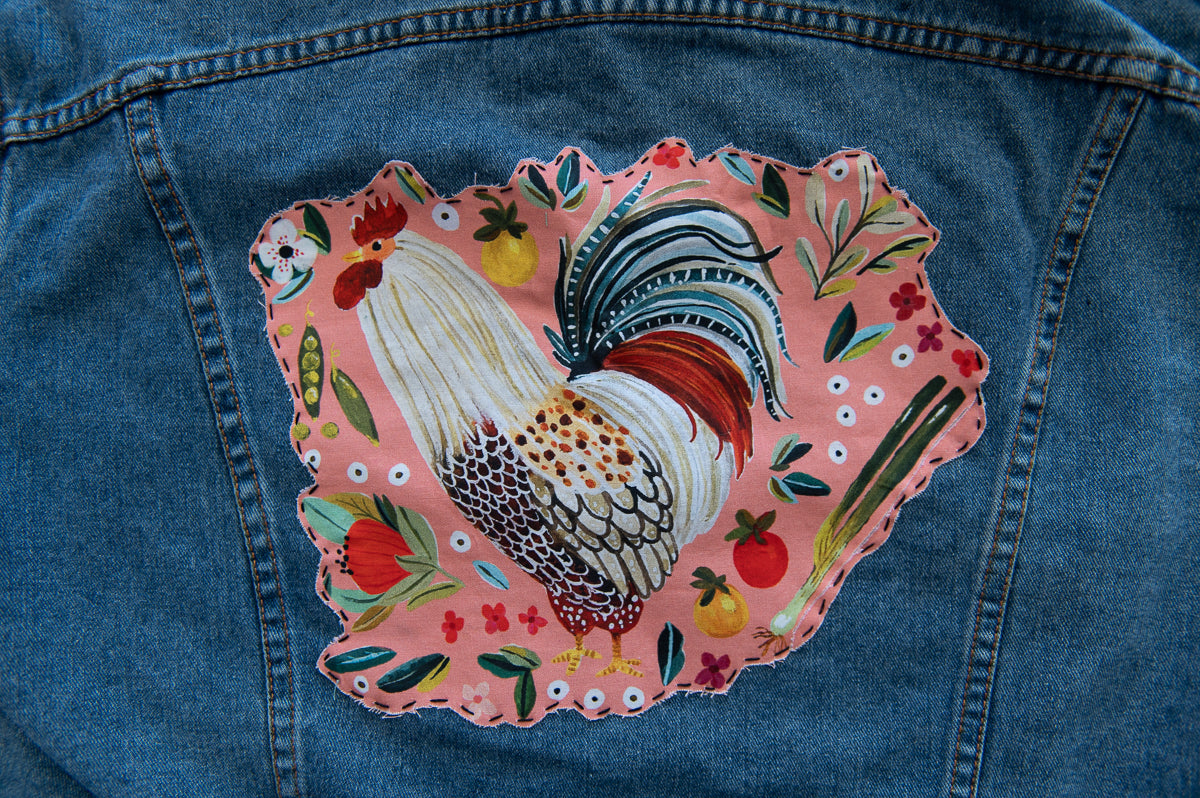 Denim jacket with a colorful rooster patch on a pink background