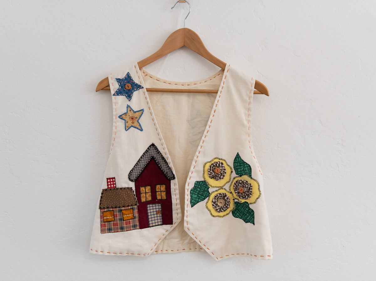 Children's vest with embroidered designs of houses and flowers on a wooden hanger against a white background