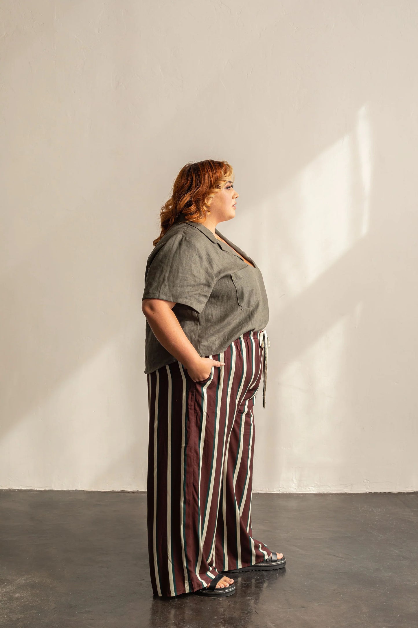 Woman wearing a gray top and striped pants standing against a plain white wall.