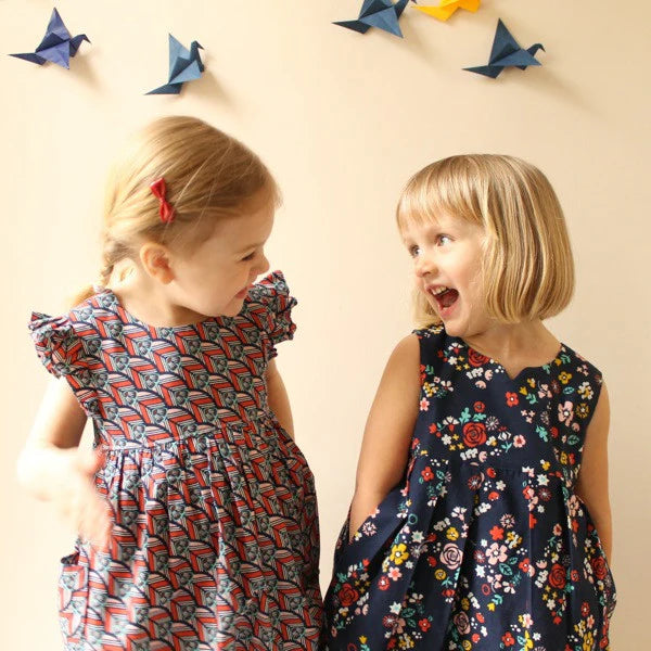 Two young girls in patterned dresses standing next to each other with colorful paper airplanes on a wall.