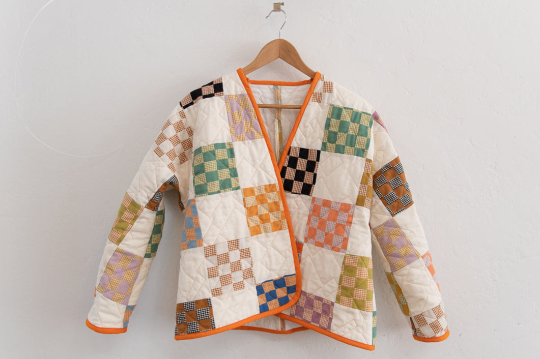 A patchwork quilt coat with bright checkered orange, green, black and orange squares on a white background, with orange bias binding