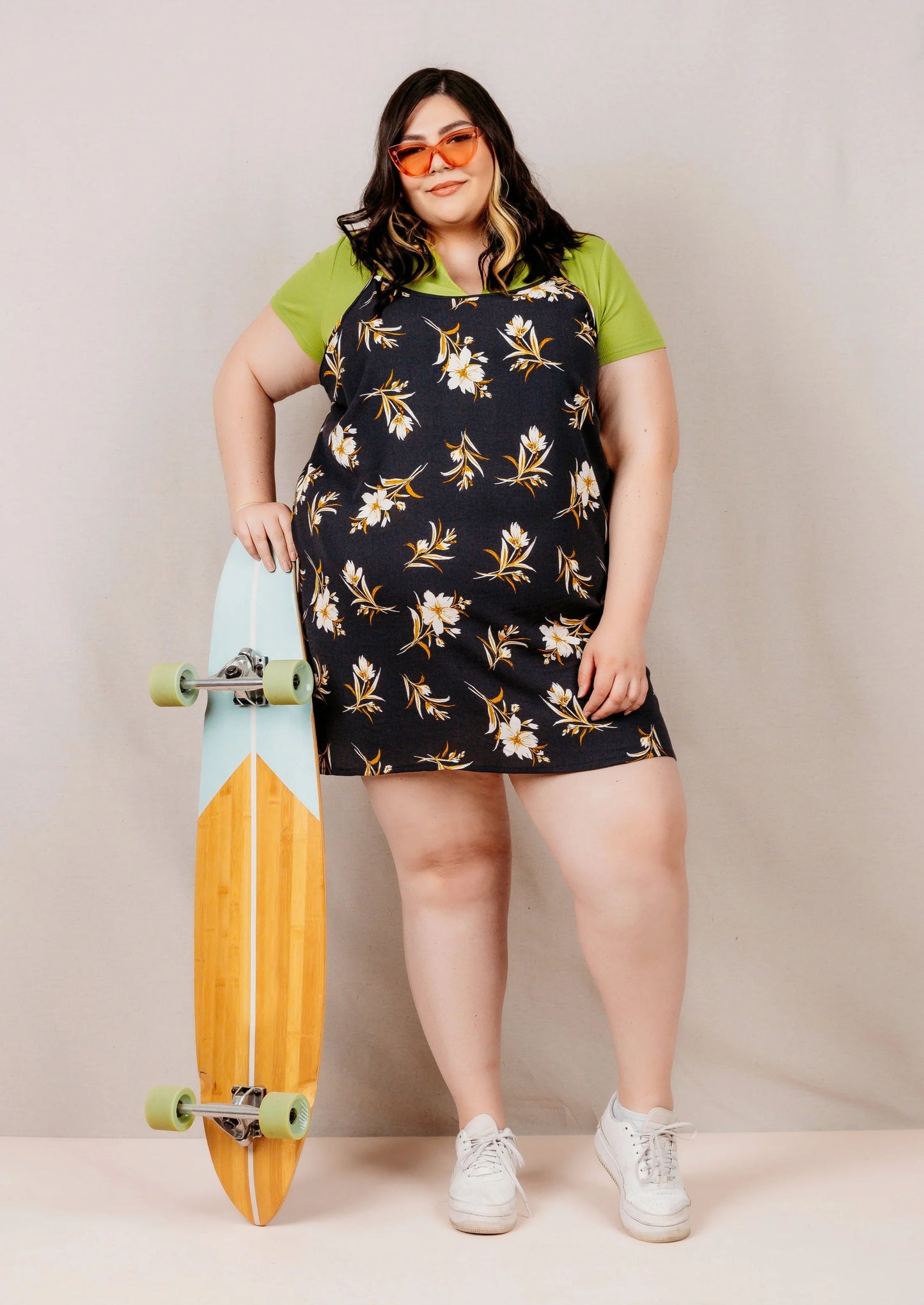 Woman holding a skateboard wearing a short floral dress and green shirt against a plain background