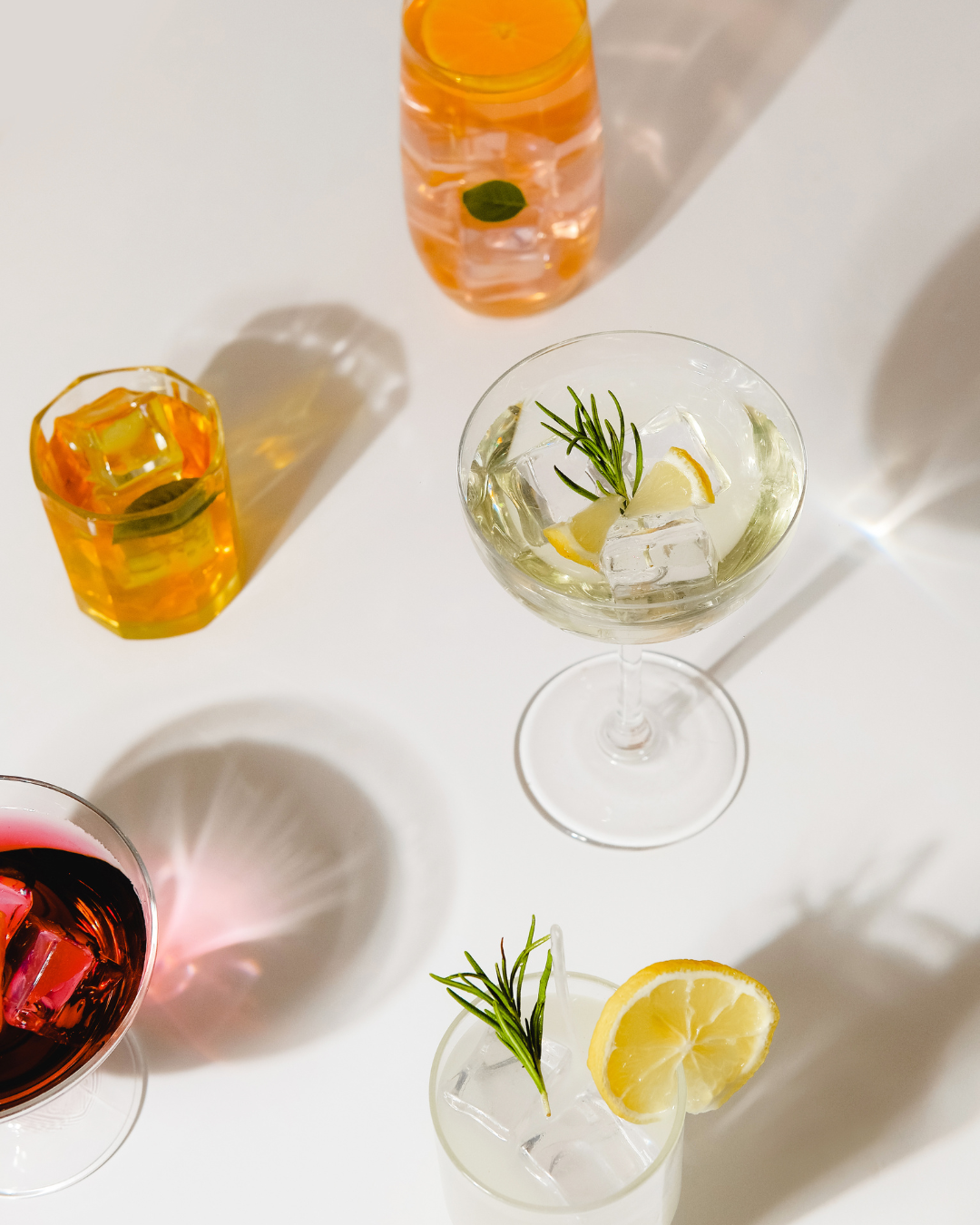 An overhead photo of various cocktails on a white surface, with one glass containing a rosemary sprig, another with a lemon slice, and others filled with orange and red liquids.