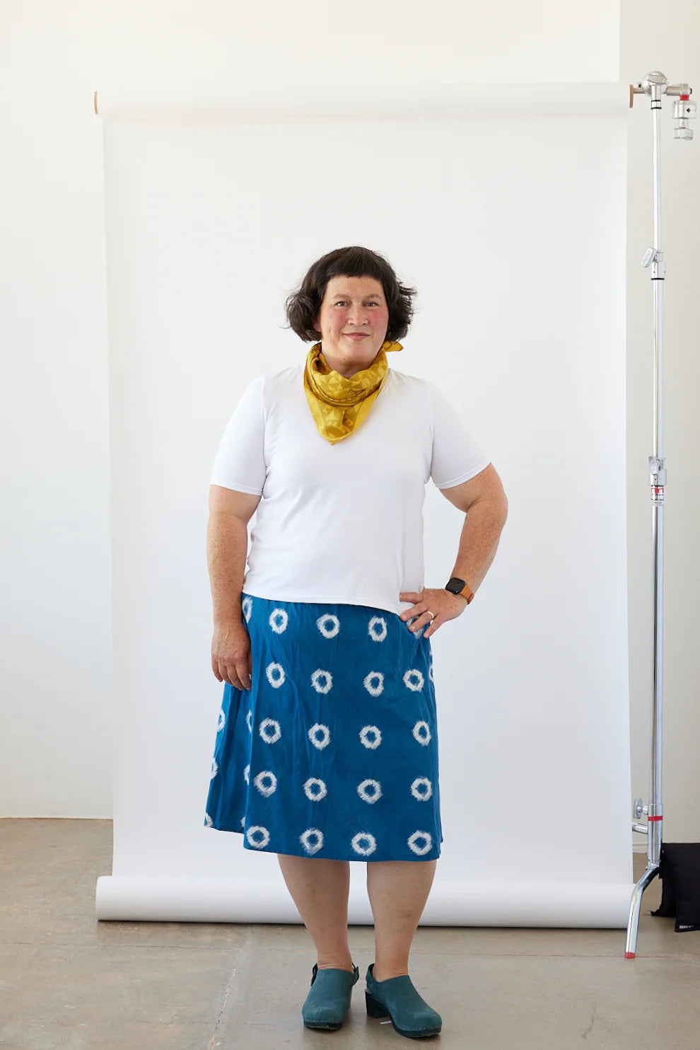 Woman wearing a white shirt, blue skirt with white patterns, and a yellow scarf against a white background.