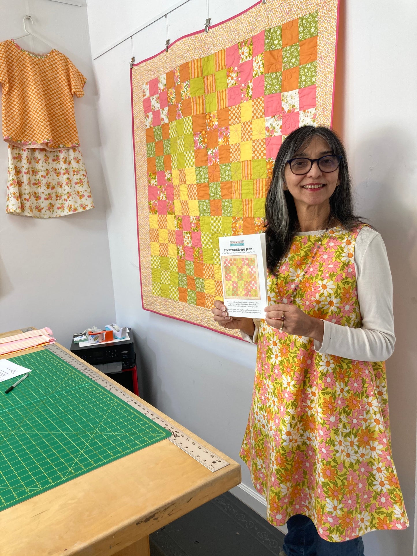 Lana holds a pattern and stands in front of a patchwork quilt