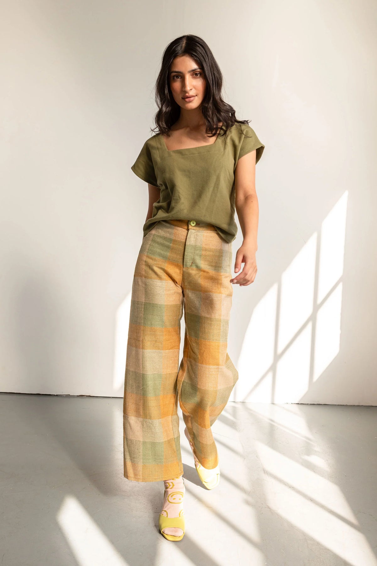 A woman modeling a loose olive green short-sleeve top and wide-leg plaid trousers in shades of yellow, orange, and green, standing in a bright studio with window light casting shadows.