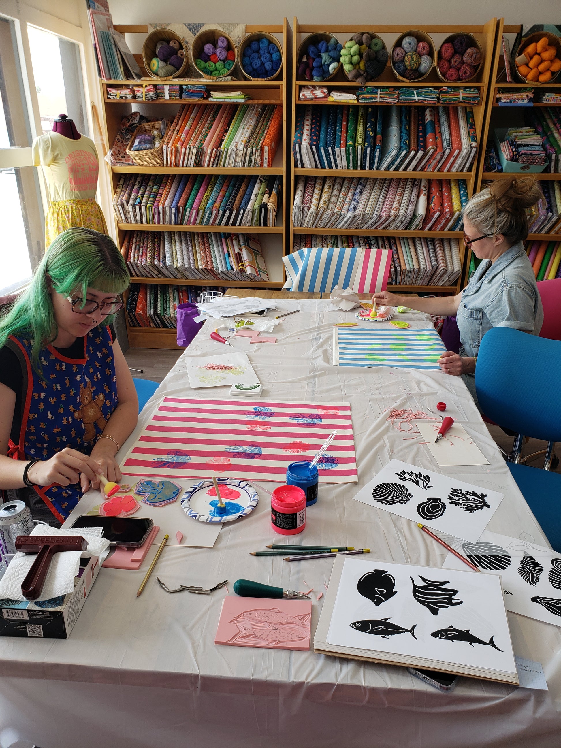 Two people working on block printing projects at a table with supplies in a room filled with fabric and craft materials.