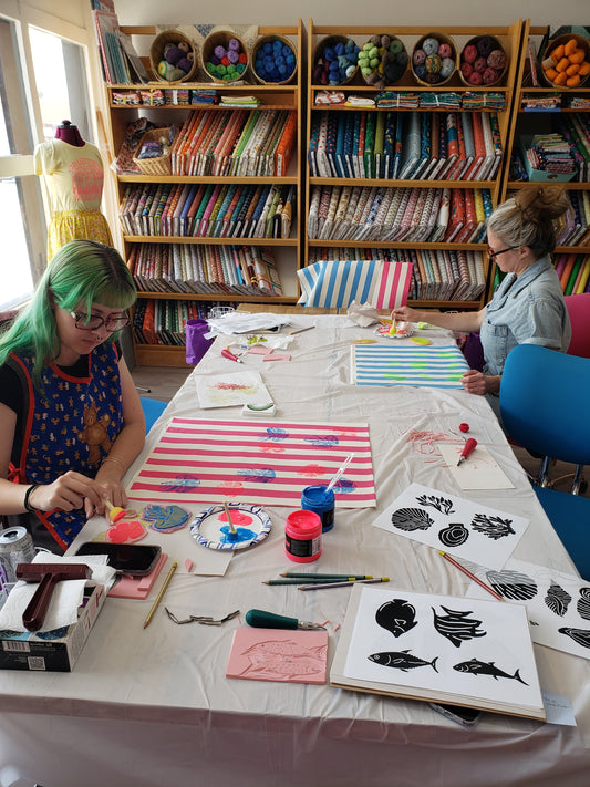Two people working on block printing projects at a table with supplies in a room filled with fabric and craft materials.