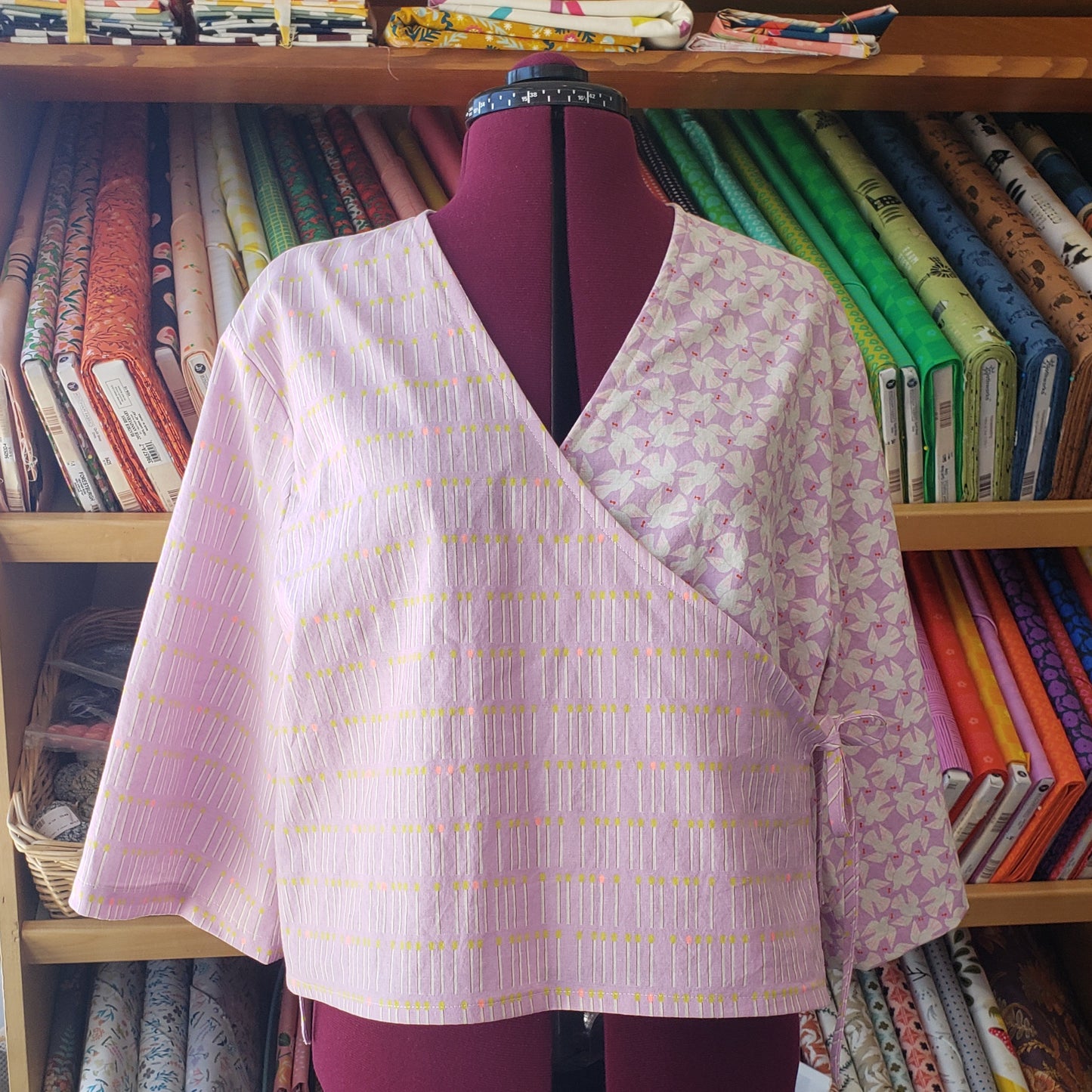 A lavender wrap-style top with a subtle floral pattern and thin yellow dotted lines, displayed on a burgundy dress form in front of shelves filled with colorful fabrics.