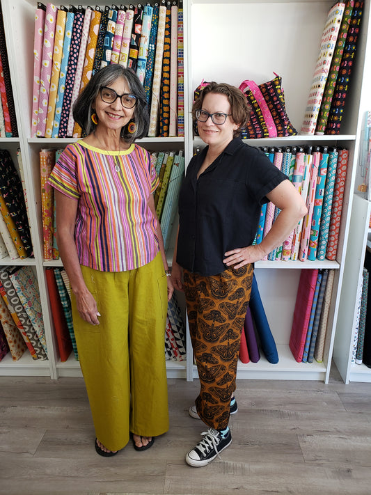 Two women standing in a fabric store showroom with shelves of colorful patterned fabrics behind them. The woman on the left wears a multicolor vertically striped short-sleeve top and mustard yellow wide-leg pants.