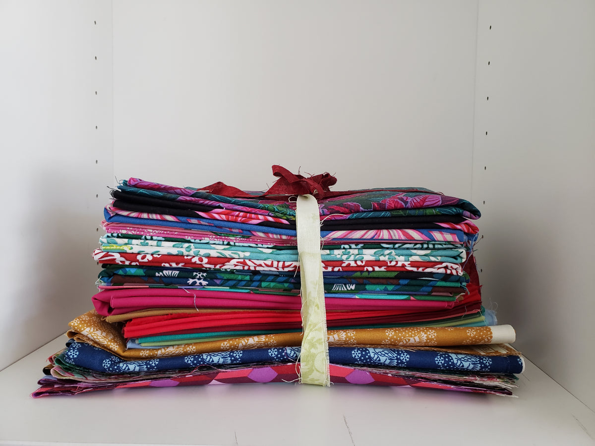 A stack of folded patterned fabric bundles tied together with a cream ribbon and red bow, displayed on a white shelf against a white wall.