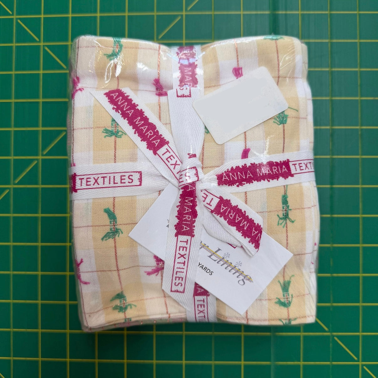 A folded piece of Anna Maria Textiles cotton fabric with a beige and white plaid pattern and green floral motifs, wrapped in clear plastic with a branded ribbon and tag, set on a green cutting mat.