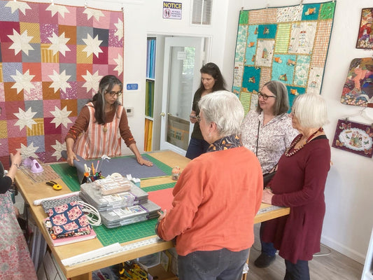 a woman at a cutting table shows a group of students how to cut fabric