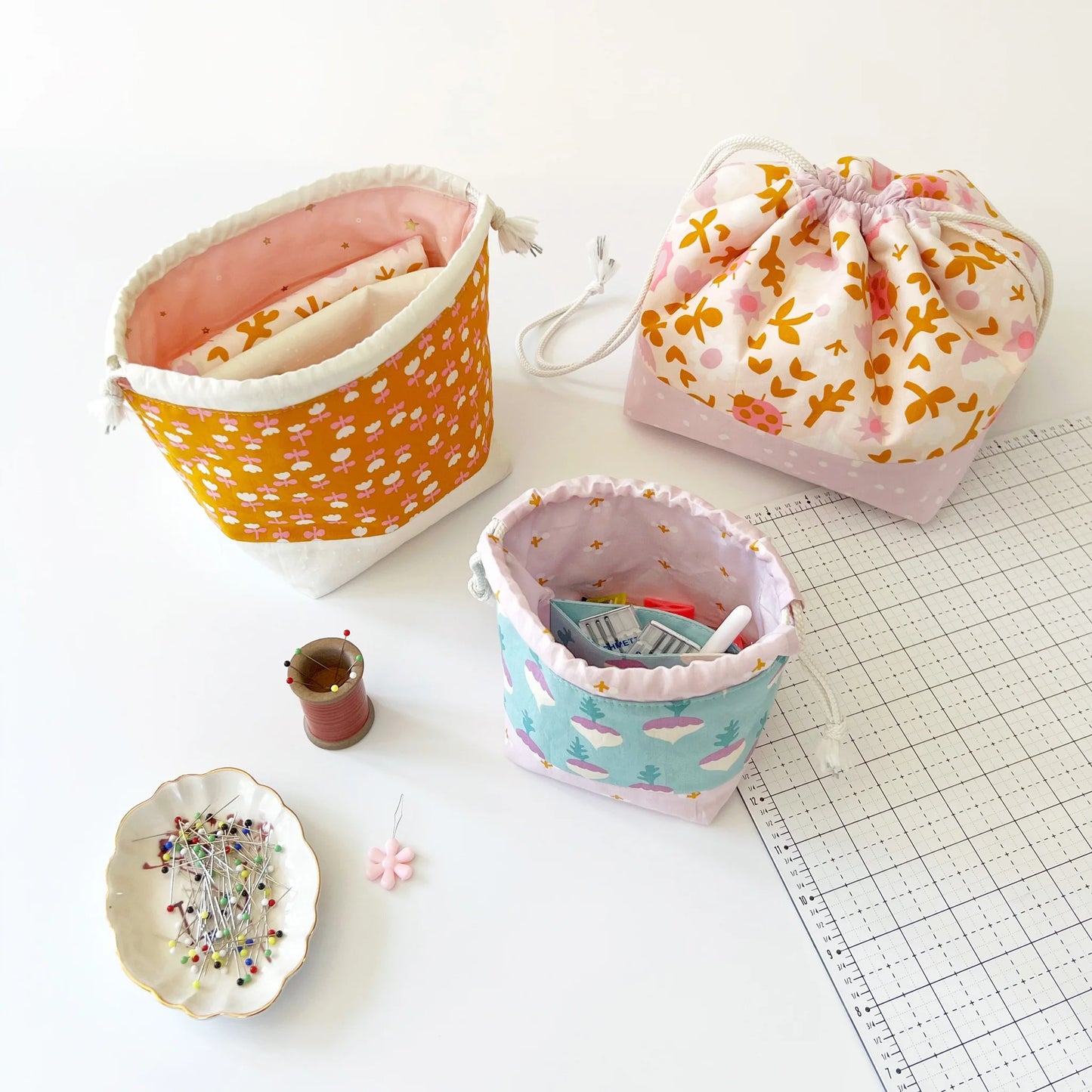 Three fabric drawstring pouches with different pastel patterns are arranged on a white surface alongside sewing props, including a pin cushion, thread spool, and cutting mat.