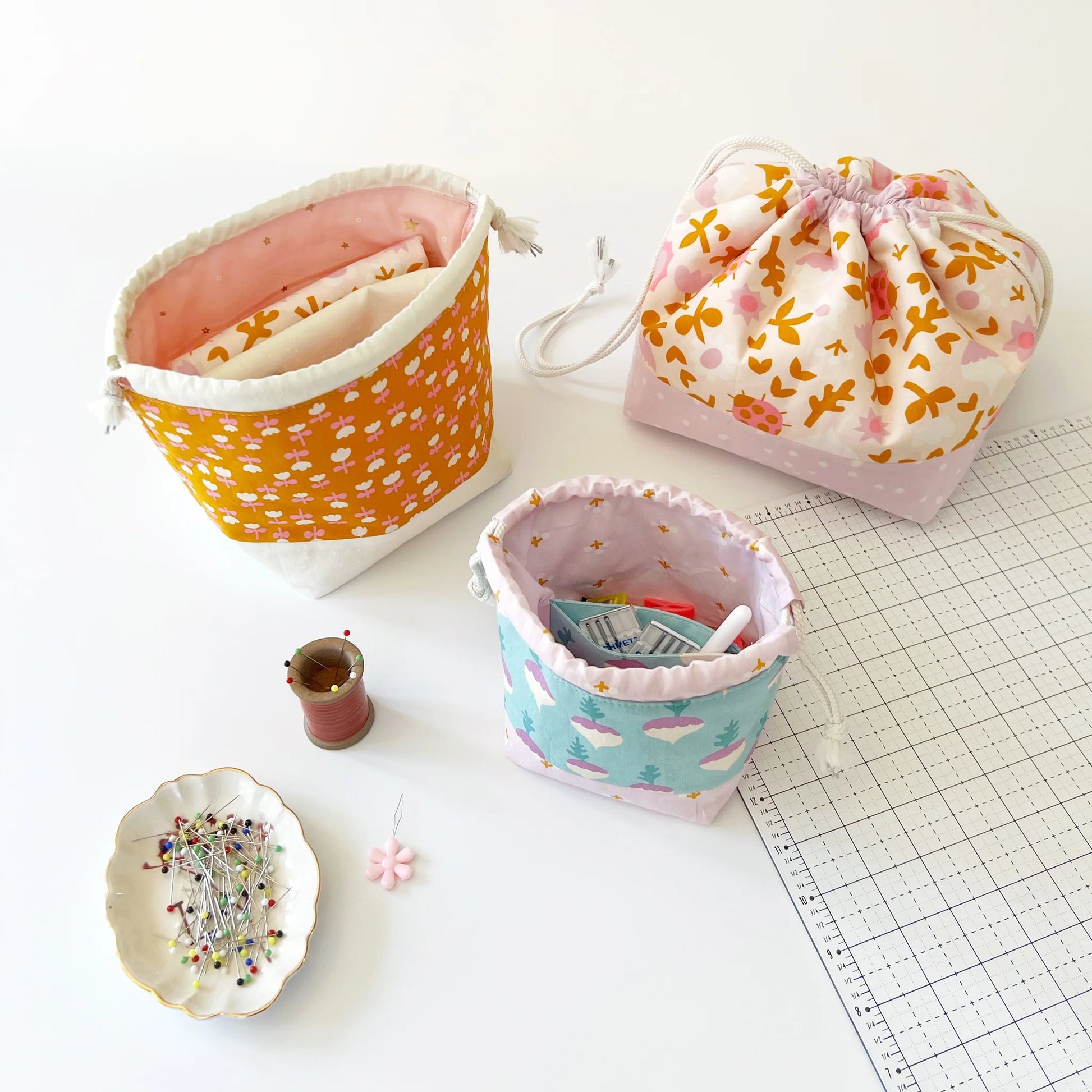 Three fabric drawstring pouches with different pastel patterns are arranged on a white surface alongside sewing props, including a pin cushion, thread spool, and cutting mat.