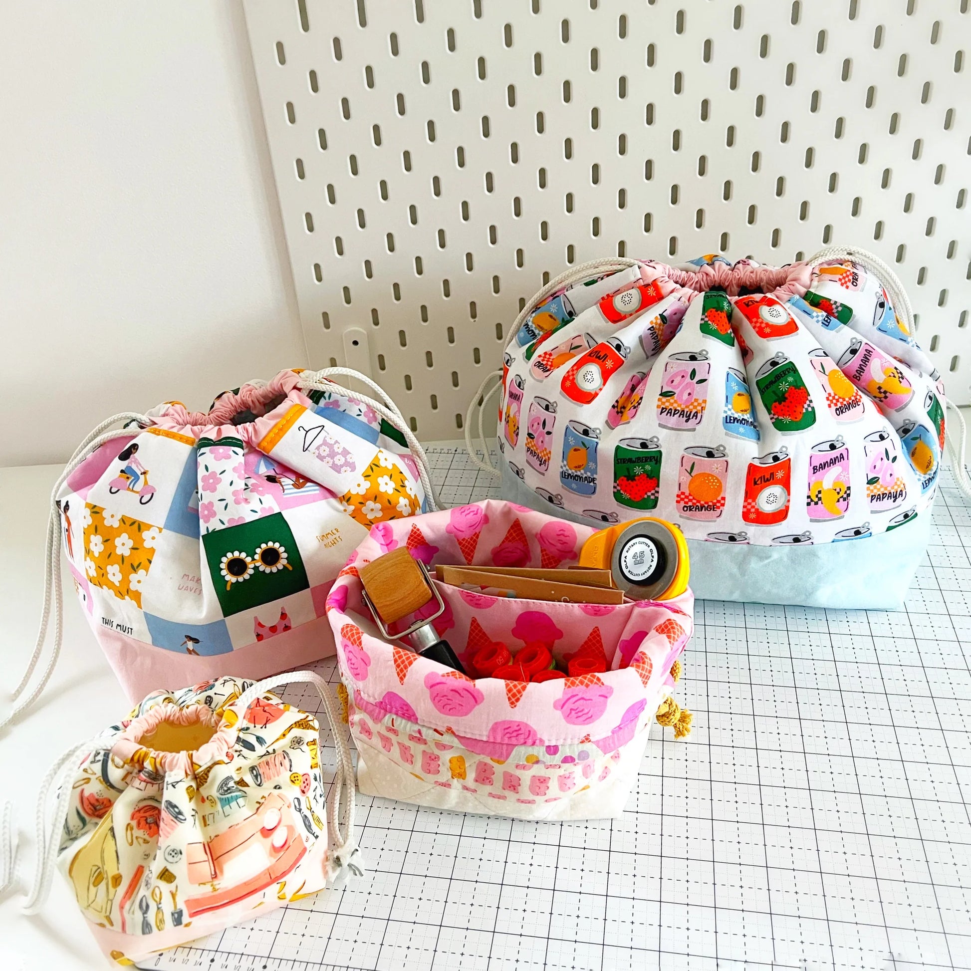 Four fabric drawstring pouches in assorted colorful prints and patterns, arranged on a white grid mat against a white pegboard background.