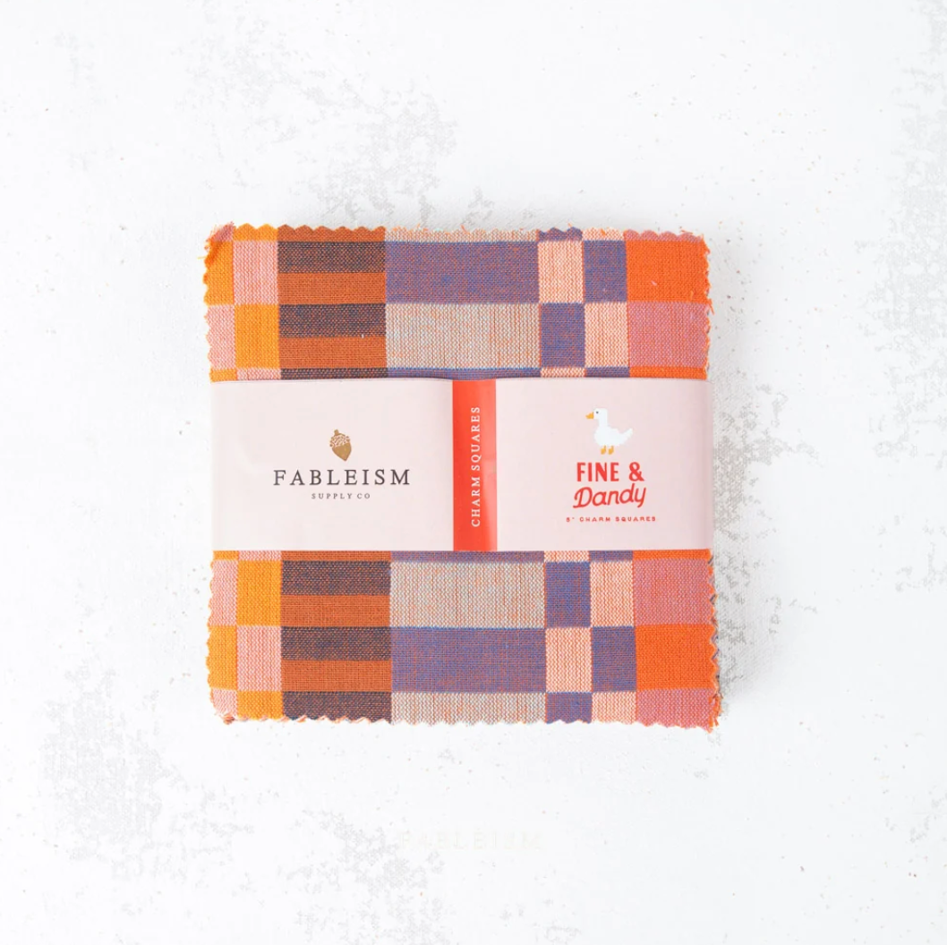 A folded bundle of Fableism Apparel fabric pieces wrapped in a pink paper band, featuring a multicolor plaid pattern in orange, purple, pink, and brown on a light textured white background.