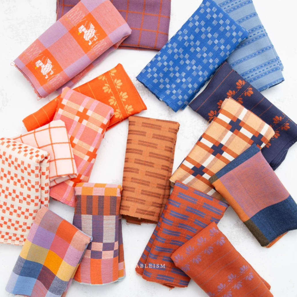 A flat-lay arrangement of multiple folded fabric handkerchiefs in various colors and patterns, including plaid, checkered, and geometric designs. The fabrics feature shades of orange, blue, pink, purple, and white, with some showing woven animal motifs.
