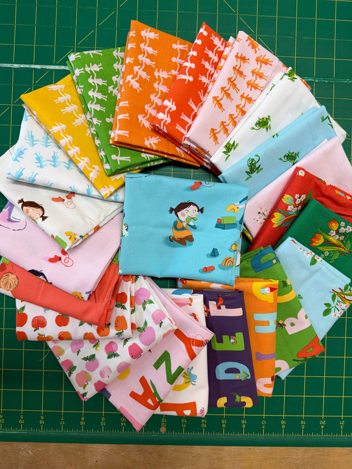 A collection of colorful fabric squares with various cartoon and pattern designs, arranged in a fan shape on a green cutting mat.