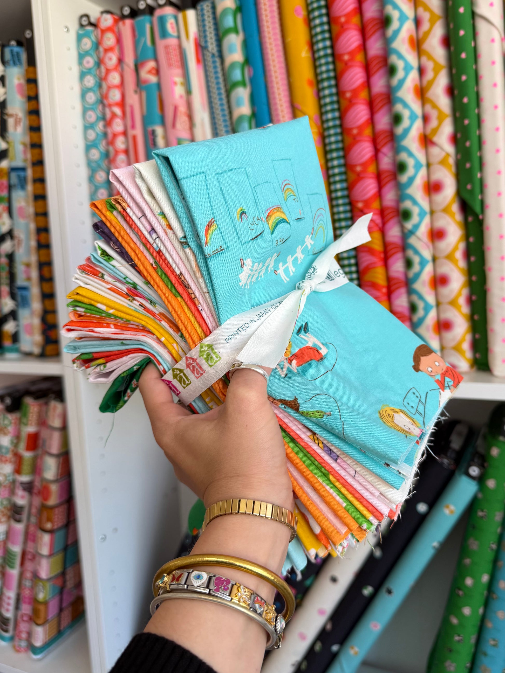 A hand holding a fanned-out selection of colorful fabric swatches, with shelves of more patterned fabrics in the background.