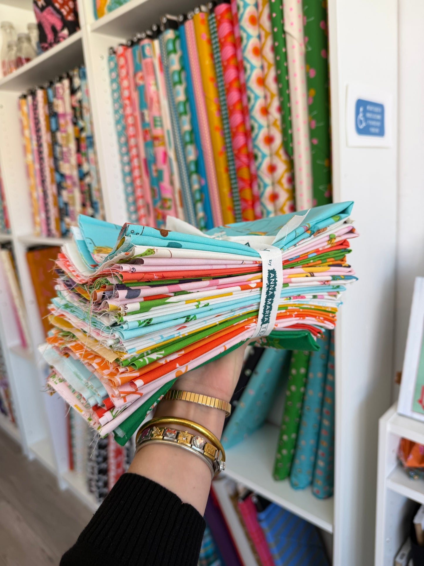 A hand holding a bundle of colorful patterned quilting fabrics, with shelves of folded fabrics in the background.