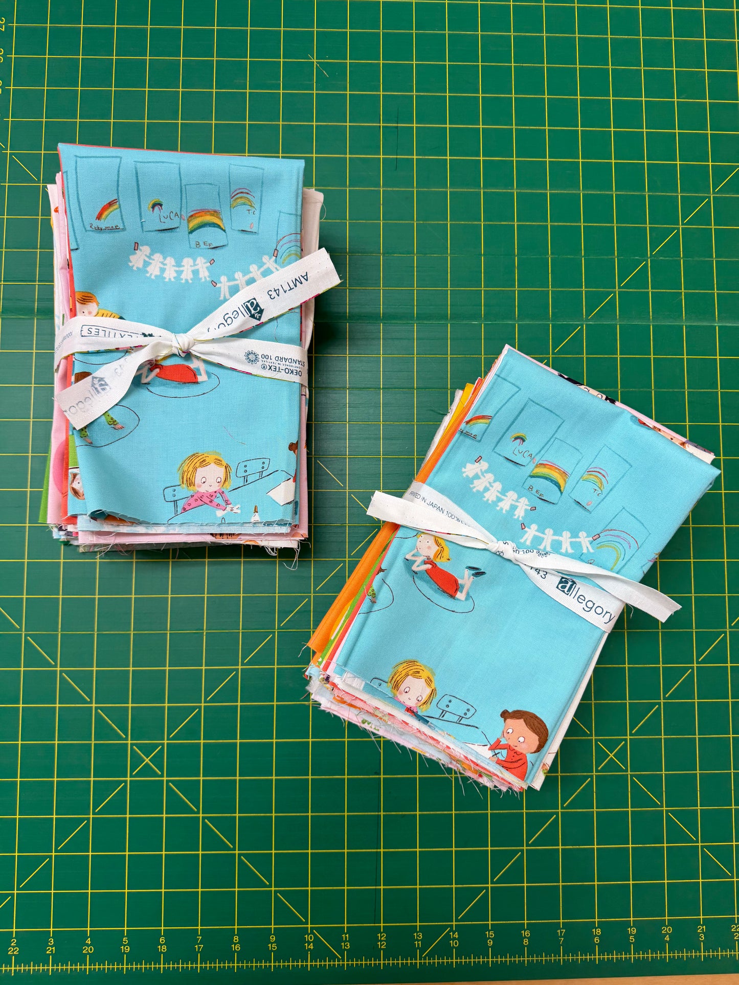 Two stacks of small fabric bundles tied with white ribbon, featuring a light blue print with cartoon children, rainbows, and stars, arranged on a green cutting mat.