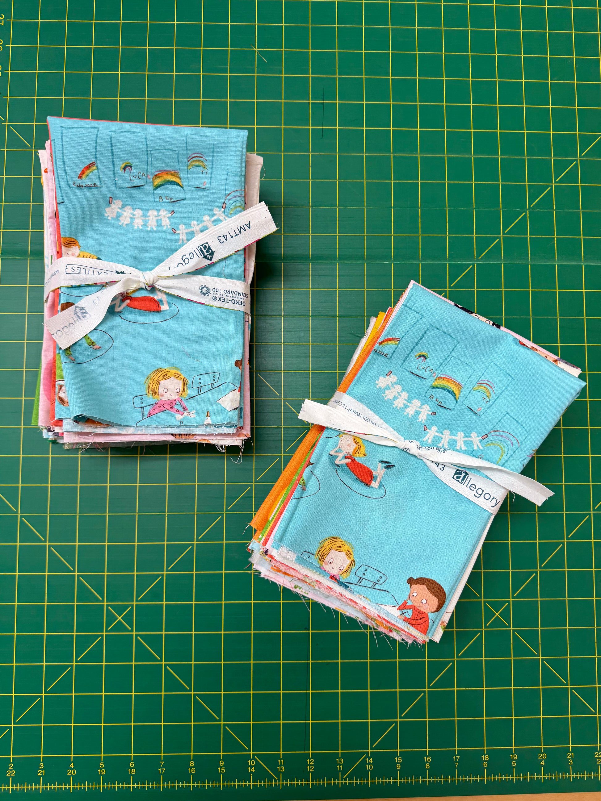 Two stacks of small fabric bundles tied with white ribbon, featuring a light blue print with cartoon children, rainbows, and stars, arranged on a green cutting mat.