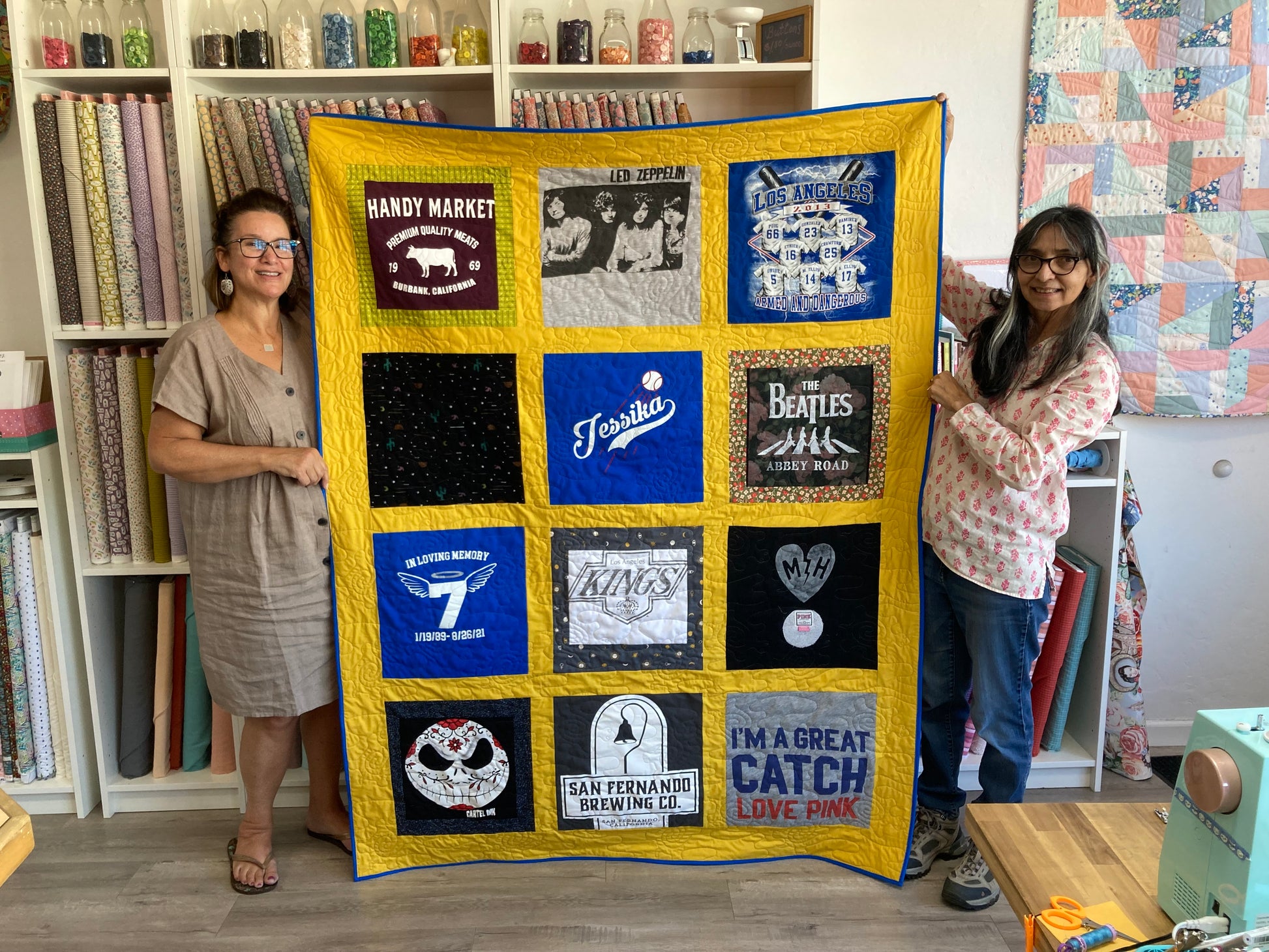 Two women holding a quilt made from various t-shirts in a room with shelves and fabric.