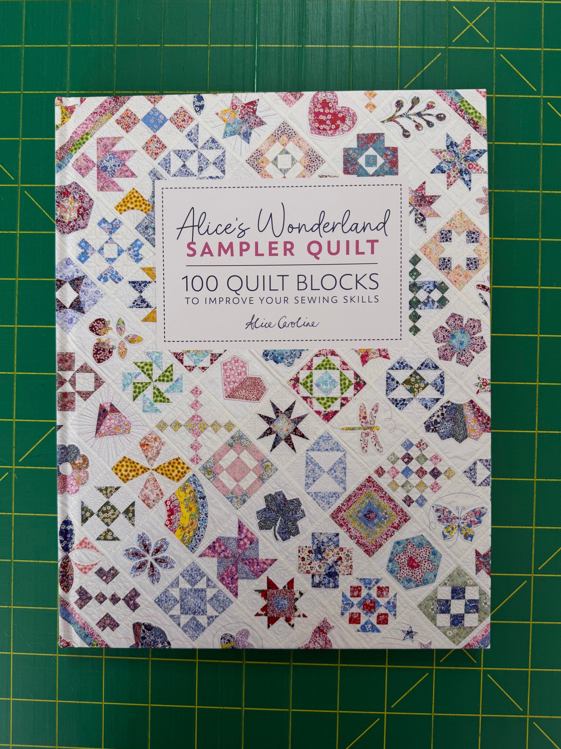 Alice's Wonderland Sampler Quilt book by Kim Carter, featuring a colorful quilt block pattern cover, placed on a green cutting mat.