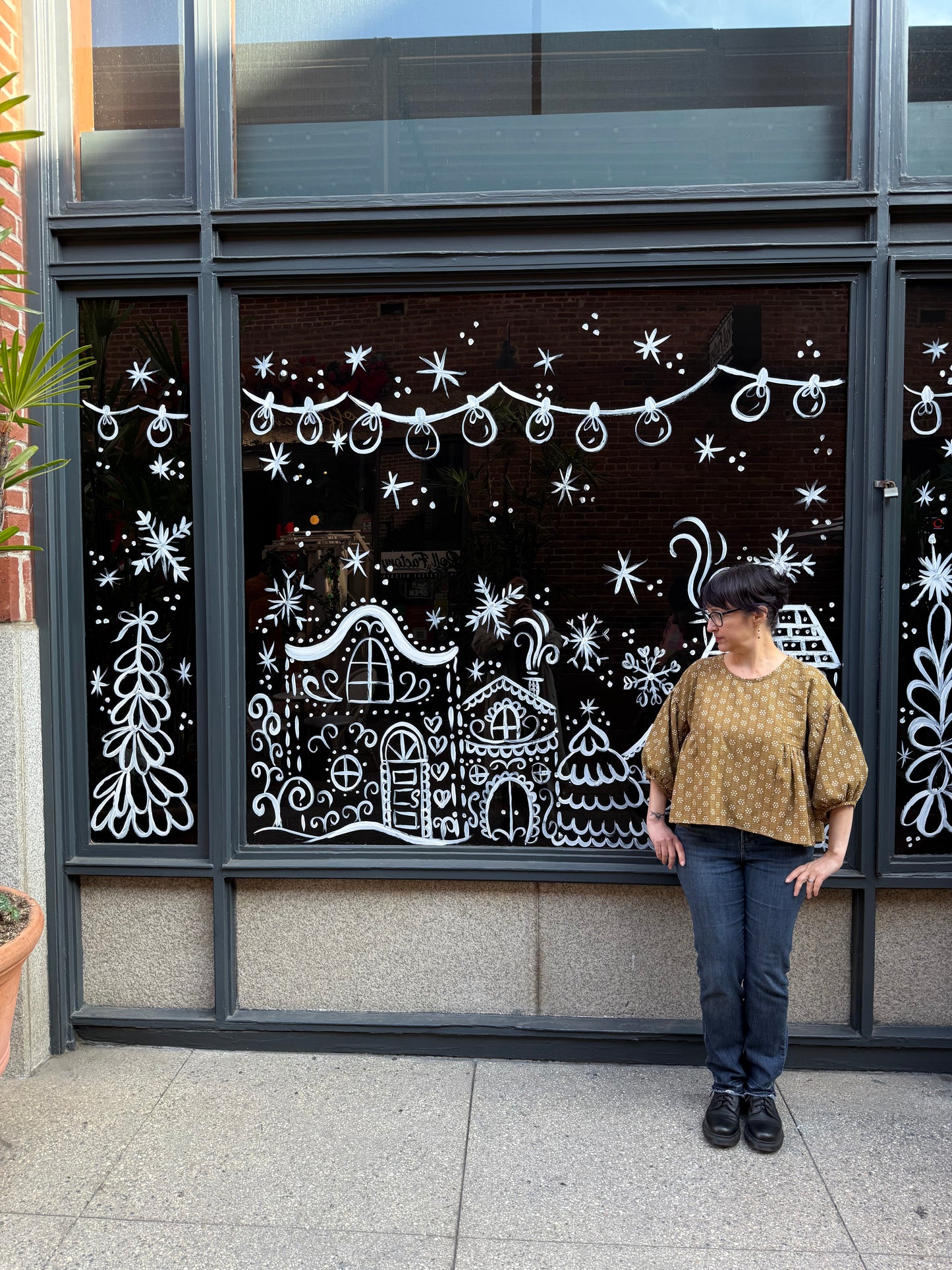 Darcy stands by a decorated window wearing jeans and a mustard colored cropped blouse with puffy sleeves.