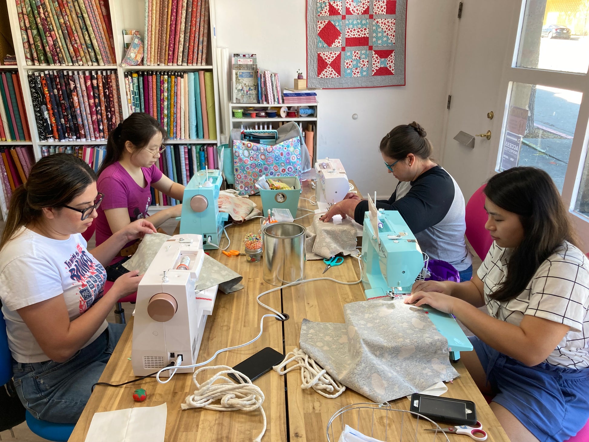4 people using sewing machines seated at a table with fabric in the background