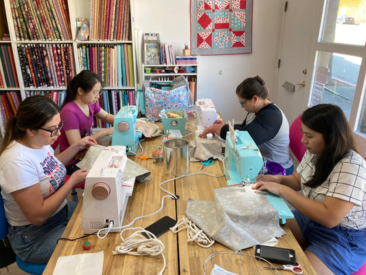 4 people using sewing machines seated at a table with fabric in the background