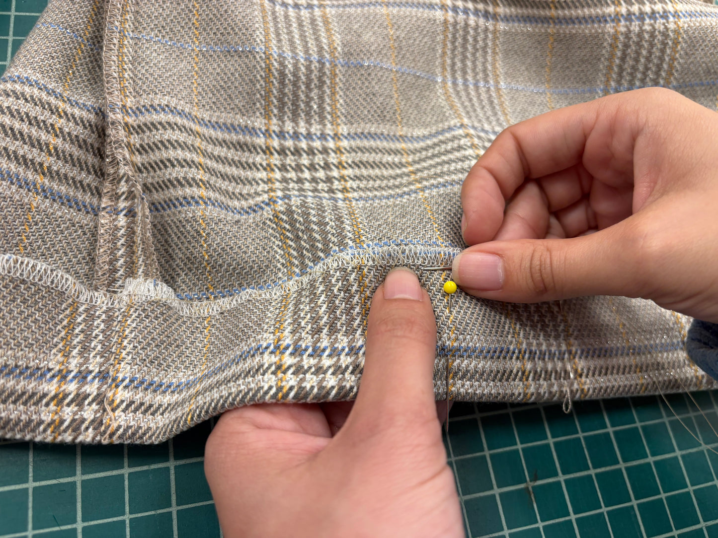 Person hand sewing a hem on plaid fabric with a needle on a green cutting mat