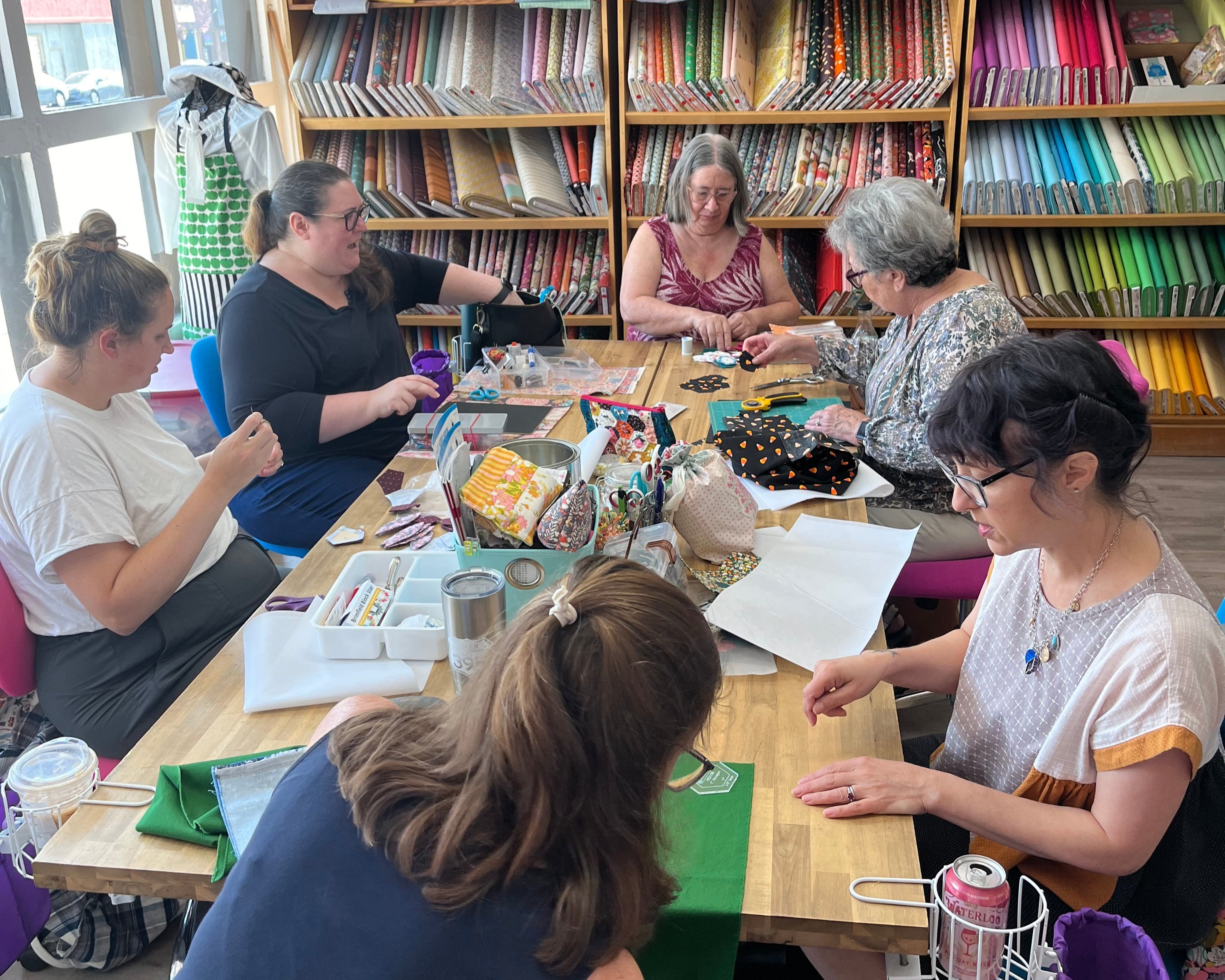 People gathered around a table learning how to sew,  in a room with shelves filled with colorful fabric.