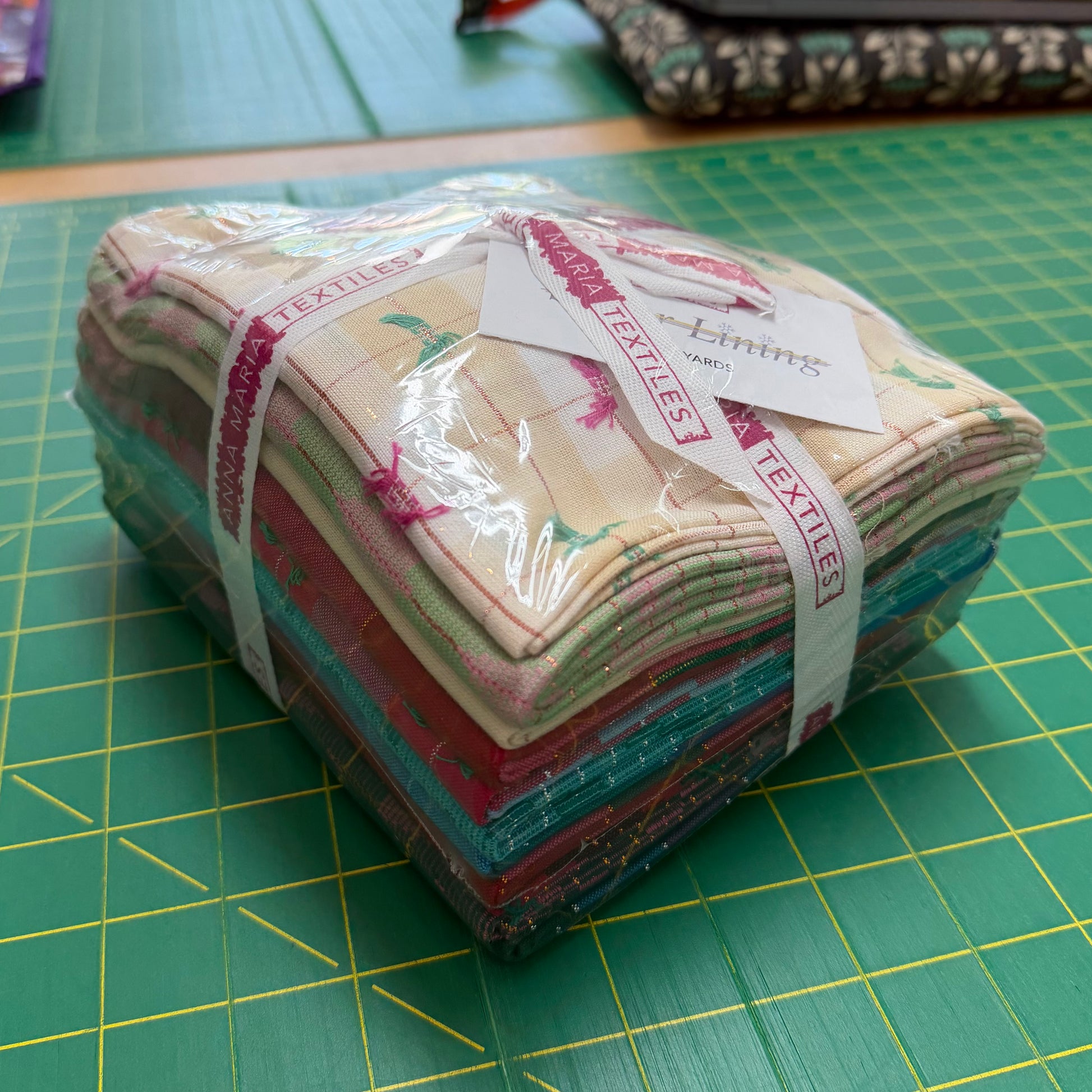 A stack of folded fabric pieces wrapped in clear plastic and tied with a white ribbon printed with the brand name 'Anna Maria Textiles', placed on a green cutting mat.