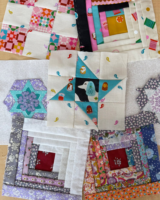 A collection of colorful handmade quilted fabric blocks arranged on a light wood surface. The blocks feature various patterns including floral, geometric, and solid colors, with one central block showing a dog illustration surrounded by teal and cream sections.