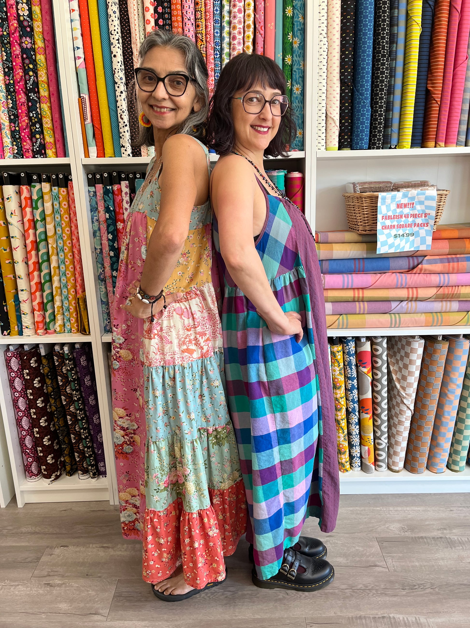 Two women wearing bright colorblock dresses standing between shelves filled with colorful fabric rolls in a store.
