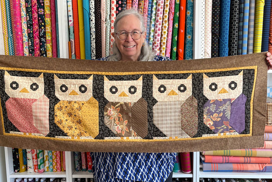 A woman holds a quilted patchwork banner featuring five patchwork owls in front of a wall of fabric bolts. 