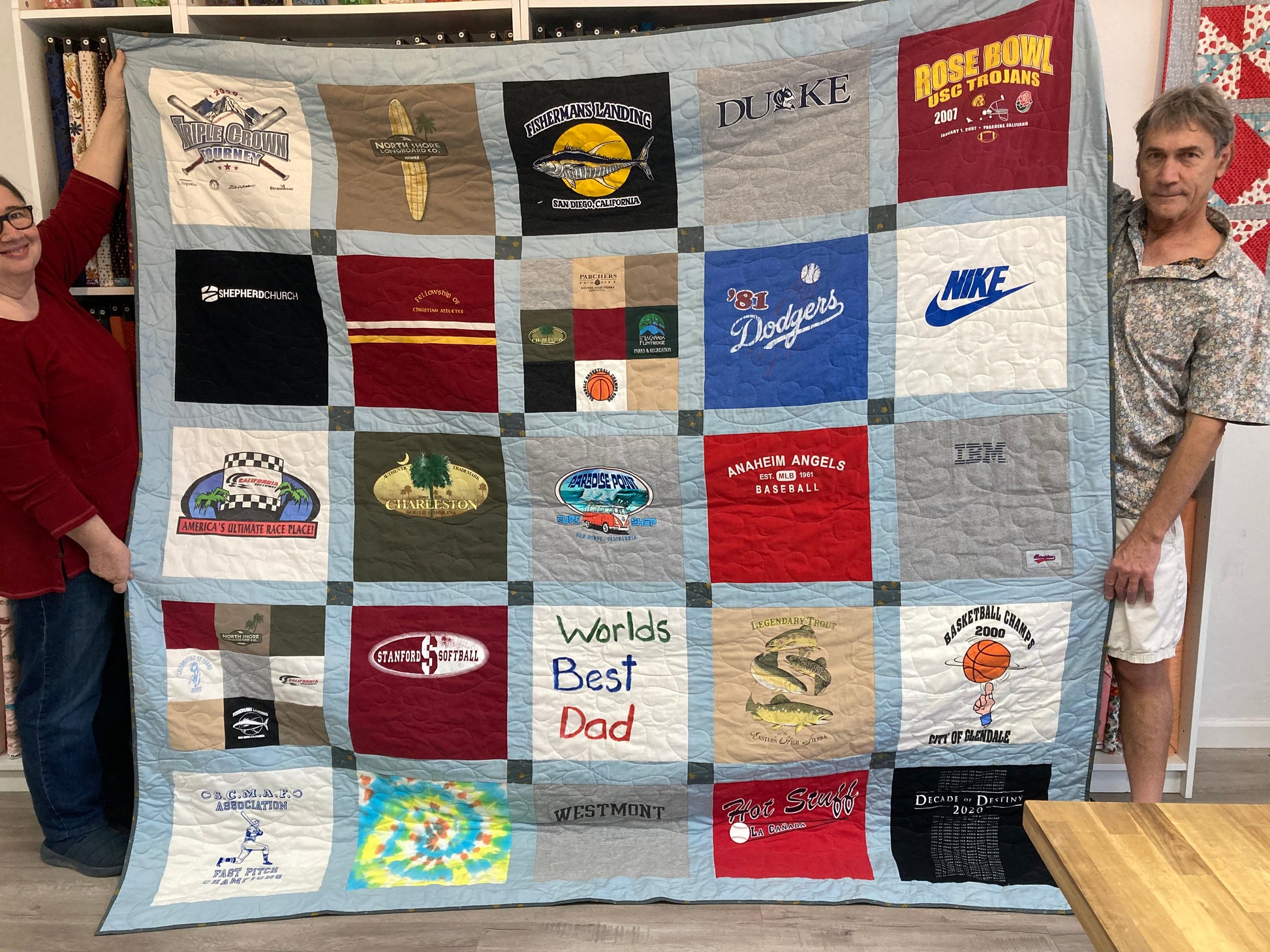 Quilt made from various t-shirts with different designs and text, held by two people.