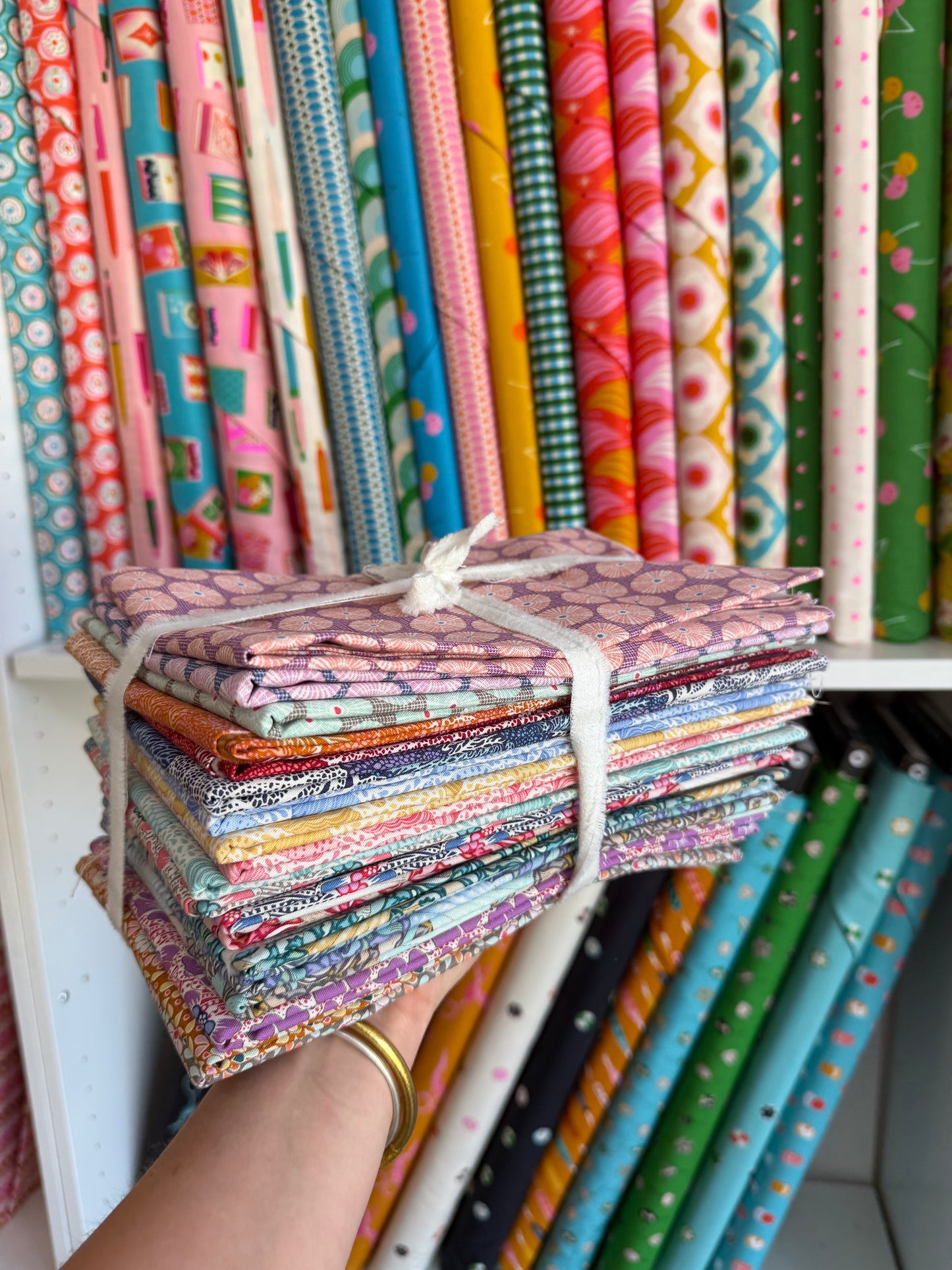 A hand holding a stack of folded patterned fabric quilts tied with white string, with shelves of colorful rolled fabrics in the background.