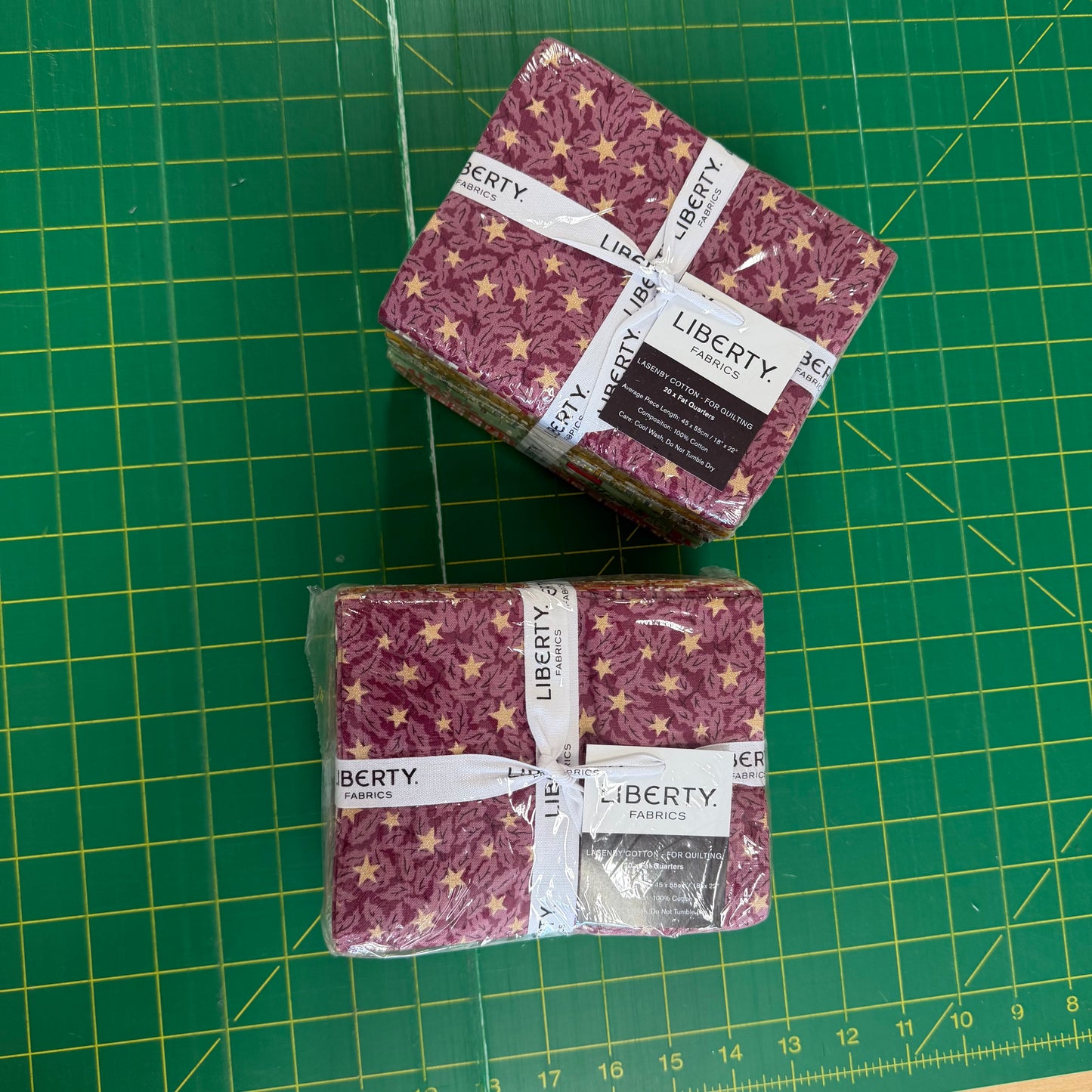 Two packaged stacks of Liberty Fabrics patterned fabric squares on a green cutting mat. The fabric has a dusty mauve/pink background with a gold star pattern.